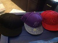 Three men's baseball caps shown side by side: black cap with black logo, purple cap with patterned brim, and red cap with embossed New York Yankees logo.