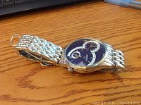 Side angle showing the band and watch face, silver-tone metal band with interlocking links, round watch face with blue dial and dual time features.