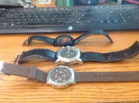 Three men's wristwatches lying horizontally on a wooden desk in front of a computer keyboard. Watches vary in strap and dial style, showing signs of wear, untested with dead batteries.