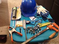 Top view of tools and safety gear arranged on a blue surface including hard hat, gloves, clamp, pipe wrench, saw, socket set, utility knife, pressure gauge, and multiple pairs of safety glasses.