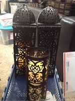 Two tall rectangular metal outside light lanterns with domed tops and intricate cut-out designs, and one smaller cylindrical lantern, all showing warm light inside.