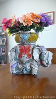 Front and side view of chalkware elephant lawn ornament with painted details, holding a pot filled with colorful artificial flowers.