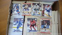 Box of assorted hockey cards with various players in team jerseys from different NHL teams, stacked and spread inside a cardboard box.