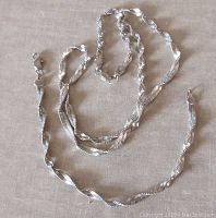 Flat lay image of twisted silver chain necklace and matching bracelet laid on neutral fabric background, showing lobster clasps and chain design details.