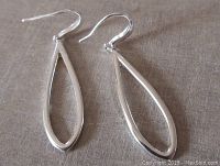 Pair of sterling silver drop earrings laid flat on a beige fabric background, showing the open elongated teardrop design and smooth polished finish