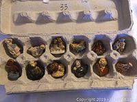 Top-down view of the 12 ceramic tea figurines arranged in a carton egg tray showing their placement and details.