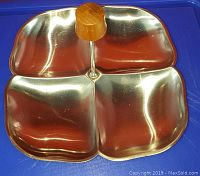 Top-down view of 4-compartment stainless steel serving tray with central teak wood handle, showing shiny polished surfaces and rounded compartment shapes.