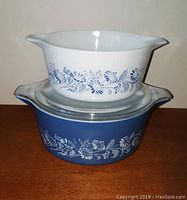 Stacked view of two Pyrex Colonial casserole bowls, smaller white on top of larger blue, showing floral patterns and double spout handles
