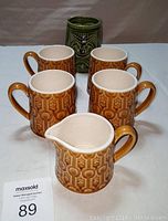 Four brown coffee mugs arranged around matching creamer with textured honeycomb pattern glaze, and an additional green ceramic coffee mug with decorative monster figure at back.