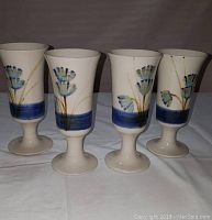 Four off-white ceramic wine glasses approximately 6.5 inches tall. Each glass features hand-painted blue and green floral designs and a blue band near the base above the stem. Glasses have wide round foot and firm stem, signed 'Tucker' on the bottom.