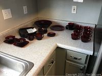 Corner countertop with arranged cranberry glass items including square plates, teacups, saucers, large bowl, ashtray, and round plate.