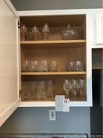 Crystal drinkware displayed on wooden shelves inside cabinet with visible range of stemware, tumblers, and covered candy dish.