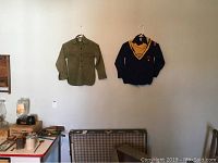 Two shirts hanging on wall, green Boy Scout shirt and navy Cub Scout sweater with neckerchief and patches, showing full front view