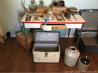 Full view showing the Lloyd metal table with red trim, drawer, and metal legs alongside the Falstaff vintage ice chest cooler with open lid, wooden-handled ice pick visible on inside top lid, and ceramic jug and crock crockery near it.