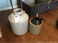 Photo showing the two crockware jugs side by side, the larger Red Wing jug with metal handle and blue wing logo marked "3", and the smaller jug with dark glazed top and cork stopper.