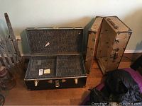 Black trunk opened showing compartmentalized interior and tray, next to tan wooden suitcase footlocker