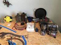 Camping supplies including canteens, cookware, first aid kit, hand warmer, tow strap, flashlight, water bag, camping stove and lantern arranged on a wooden surface.