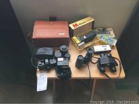 Pentax 110 camera, four lenses, flash unit, Minolta 16 PS camera box and accessories, and two binoculars on a wooden table.