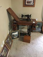 Full view of Singer treadle sewing machine in wooden cabinet with drawers and crochet box on floor nearby and framed pictures leaning against wall.