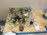Wide shot showing over 50 assorted shot glasses of varied sizes and designs arranged on a wooden surface.