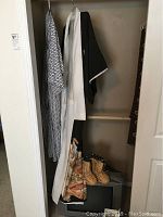 Photo showing a hanging black kimono with white collar and intricate gold and white landscape designs along the bottom, a blue and white dressing gown, and a pair of woven reed children's boots placed below them.