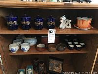 Porcelain sake teapot with blue design alongside five matching sake cups on a wooden shelf.