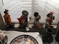 Four Japanese dolls on stands displayed in front of window blinds, dressed in traditional attire and standing on black bases.