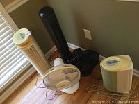 Five assorted fans together on the floor near a corner, showing beige and black tower fans, small box fan, and circular fan.