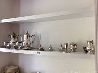 Wide view of shelf showing full tea and coffee set including multiple pots, sugar bowl, butter dish, candle holder, glass goblets, and tray
