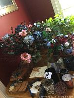 A large cluster of silk plants and flowers in brass pots, showing pink, blue, green, and red artificial flora on a wooden surface.