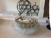 Clear glass footed punch bowl with 19 matching glass cups hanging from rim. Two black metal wine racks behind it.