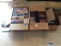 Three boxes of assorted VHS movies, music CDs, and video games shown on carpet in front of stone wall.