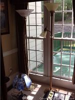 Photo showing two tall brass floor lamps with cream-colored glass shades, one with additional smaller adjustable lamp arm, and a blue and white dolphin decorative lamp on the floor nearby.