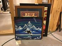 Three framed art pieces stacked leaning against a wooden post in basement. One piece features an orca whale jumping in front of snow-covered mountains and an aurora.