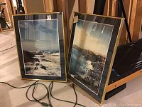 Two framed coastal ocean prints leaning against wall, showing waves and rocks.