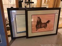 Two framed artworks leaning against a wall in a basement, showing the entirety of both pieces.