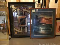 Two framed prints side by side on floor. Left is abstract geometric print; right is tropical sunset print.
