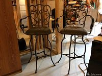 Pair of metal swivel bar chairs with cushioned seats seen from front and side angles, showing wrought iron backs with scroll and heart designs, curved armrests, and thin legs with footrests.