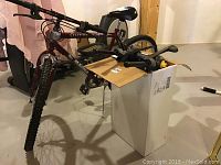 Side front angle of Manga Outreach 20 inch maroon mountain bike with front suspension and visible chipped plastic on sprocket, next to a box containing Graber 2 bike rack components.
