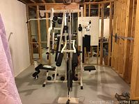 Wide view of white BMI 9800 multi-functional gym machine with padded seats and rollers in basement room without finishes