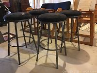 Four metal bar stools arranged in a cluster showing the black fabric cushioned tops and the metal legs with circular footrests.