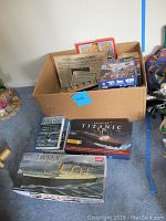 View of box containing newspaper, books, puzzles, and model kits, with three boxed model kits on floor in front.
