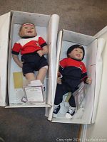 Two dolls in open white boxes showing full bodies and certificates at feet