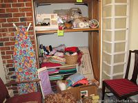 Wide shot showing ironing board with floral cover, plastic storage tower, folding table with fabrics, baskets, and sewing machine box