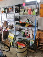 Metal shelving unit holding various bags, boxes, and containers of garden and household supplies. Collapsible fabric bin filled with bags of seeds and chemicals beneath shelves. Door mats leaning against shelving with Quikrete Walk Maker package visible.