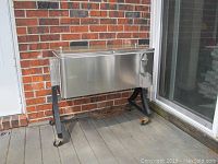 Full view of a stainless steel outdoor beverage cooler on a black stand with wheels placed outside a brick wall with glass door nearby.