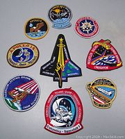 Nine vintage NASA mission patches laid out showing front designs of various Apollo and Shuttle missions, detailed embroidery colors and designs including Eagle, space shuttle, and astronaut visuals.