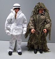 Two 12-inch GI Joe figures standing, one in white winter outfit with sunglasses, the other in brown jumpsuit with camo ghillie suit.