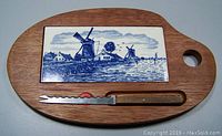 Oval wooden cheese board with blue and white Delft tile inset and serrated cheese knife with wooden handle placed below the tile.