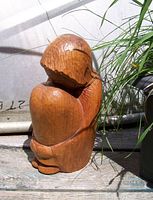Side angled view of a handcarved wooden figure depicting a seated person with short hair, hugging knees, on an outdoor wooden surface with grass nearby.